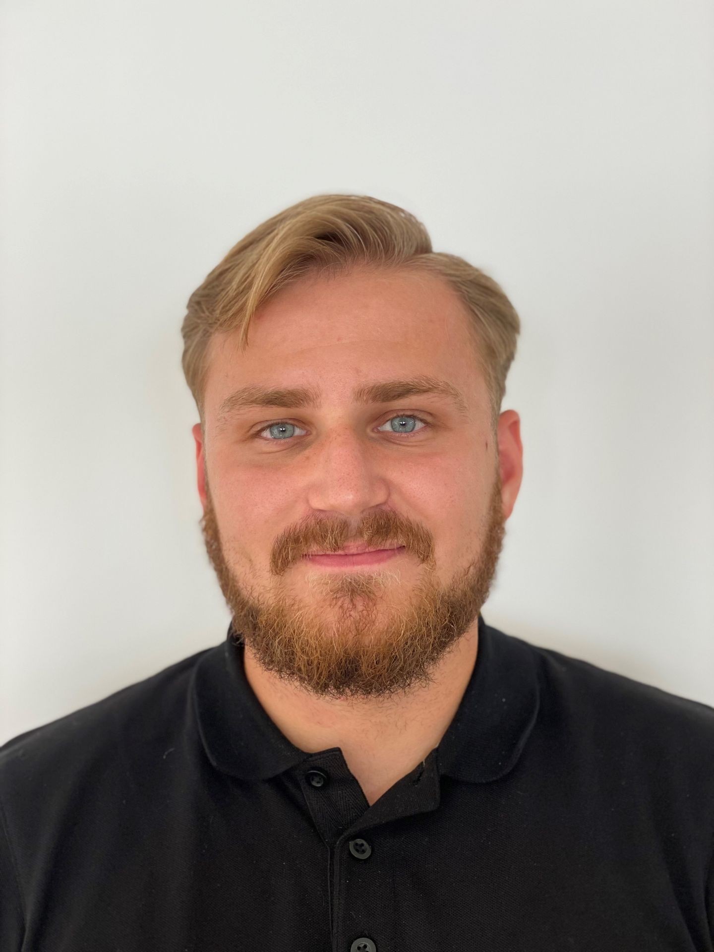 Man with beard and short blonde hair wearing a black polo shirt against a plain white background.