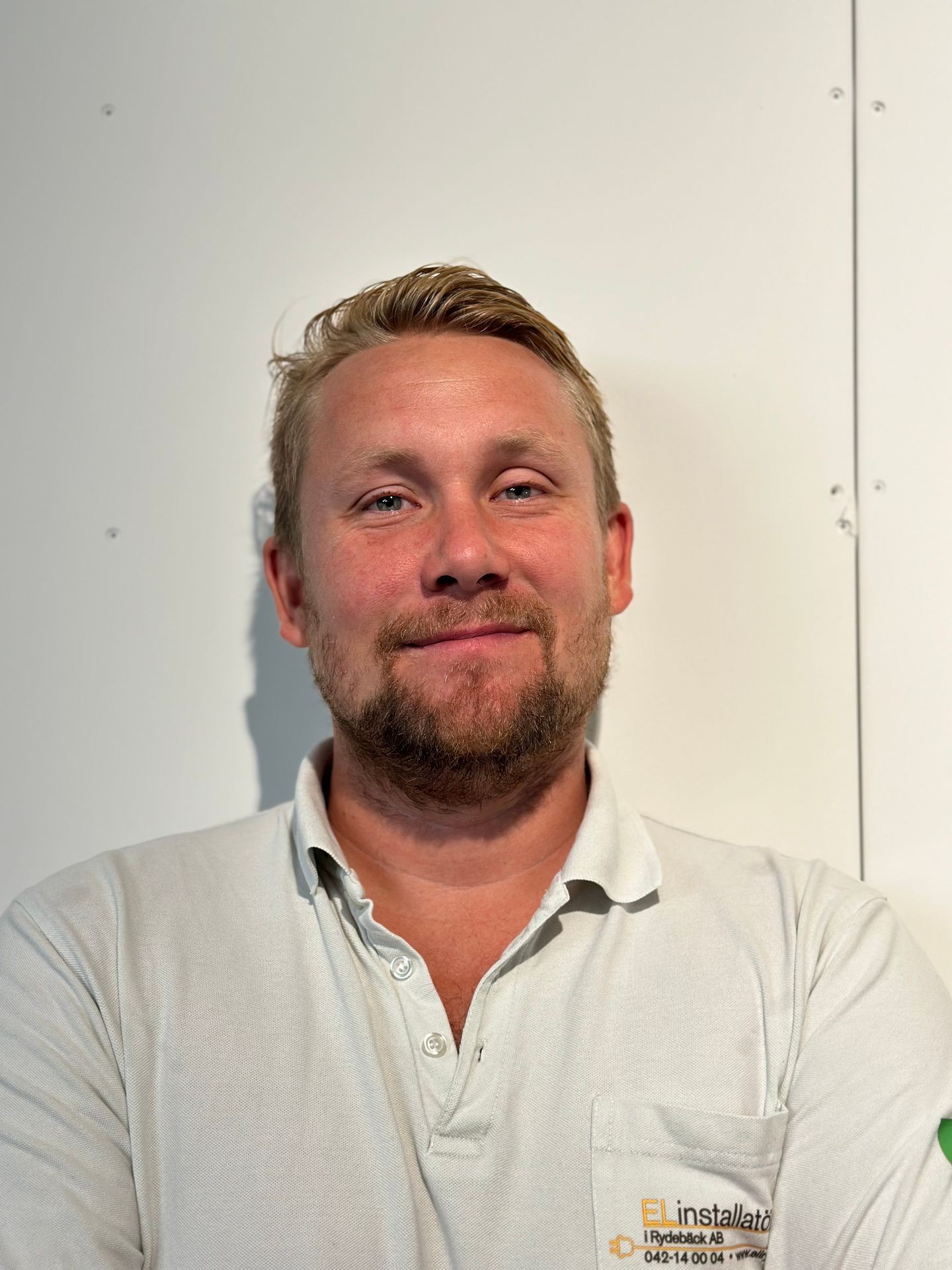 Person wearing a white polo shirt with a logo and folded arms standing against a white wall.