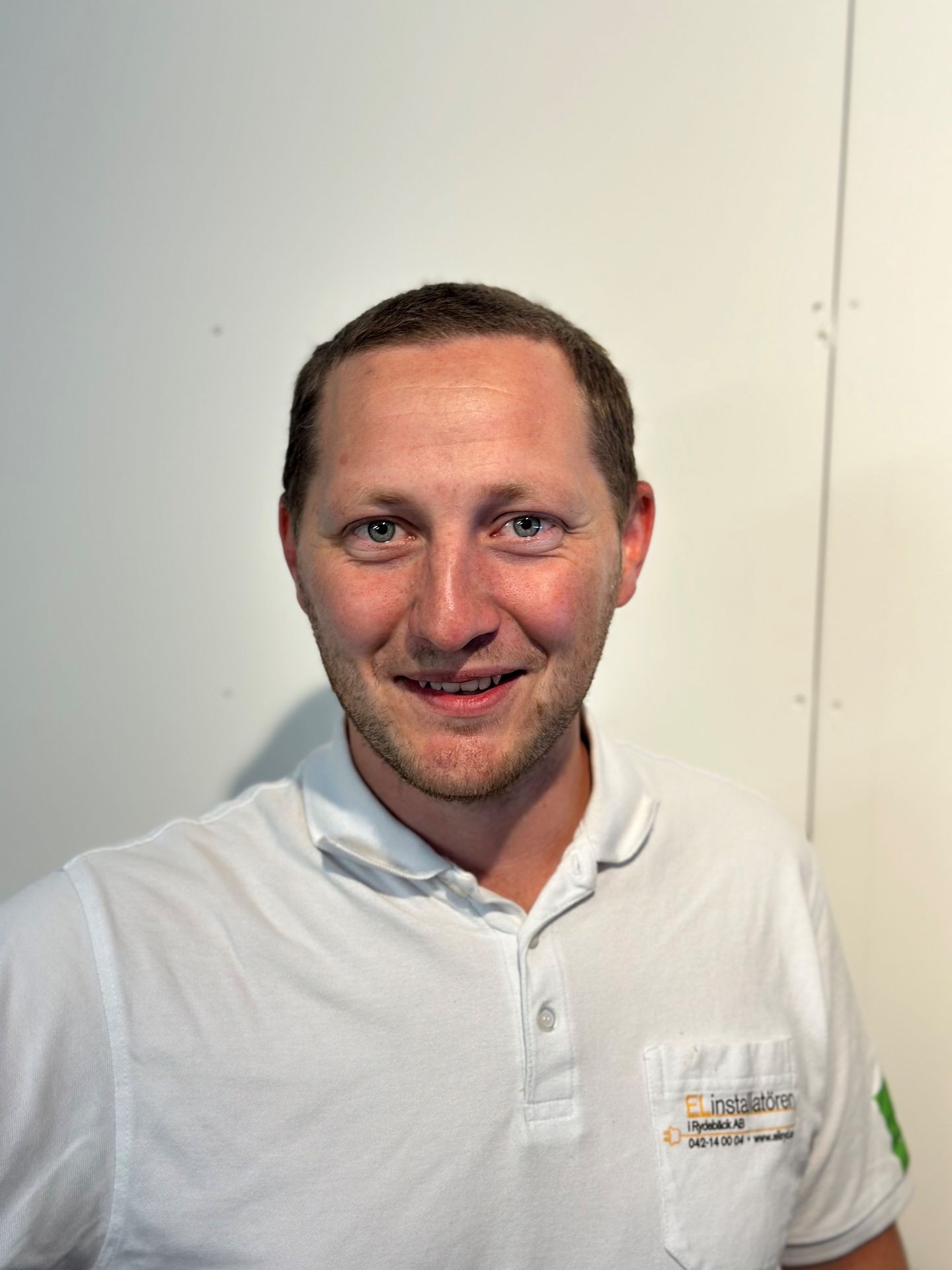 Man in a white polo shirt with a company logo, smiling and standing against a plain background.